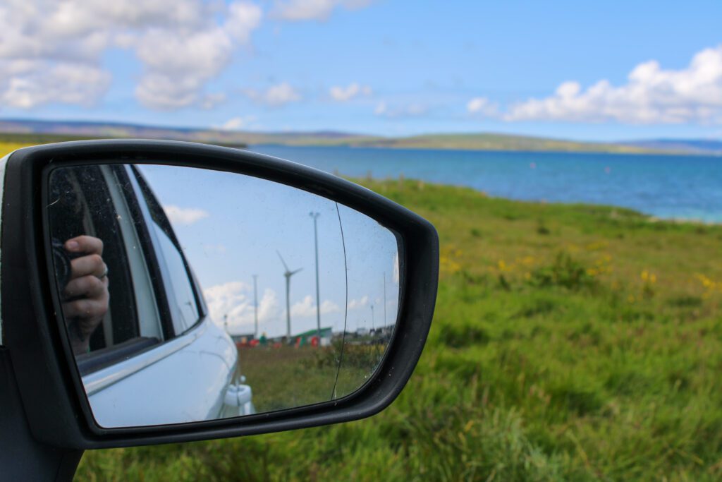 Side mirror reflection showing wind turbines and a hand holding a camera, with grassy fields and blue water in the background at Hatston, Kirkwall.