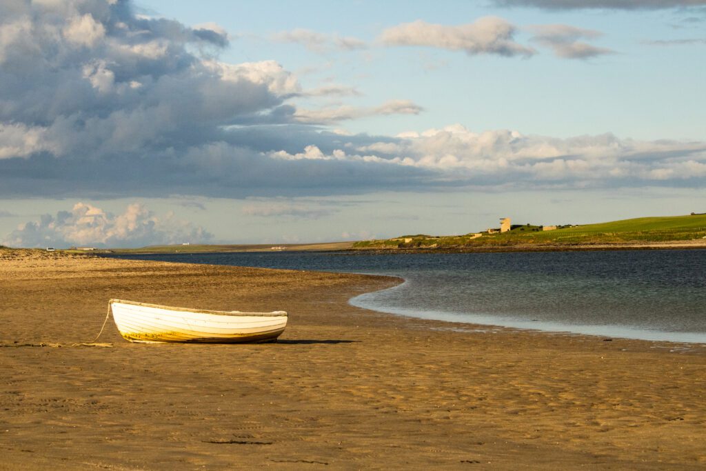 A white row boat on the sandy shore at Glimps Holm near the third Churchill Barrier in Orkney, with calm blue-green water and dramatic skies in the background